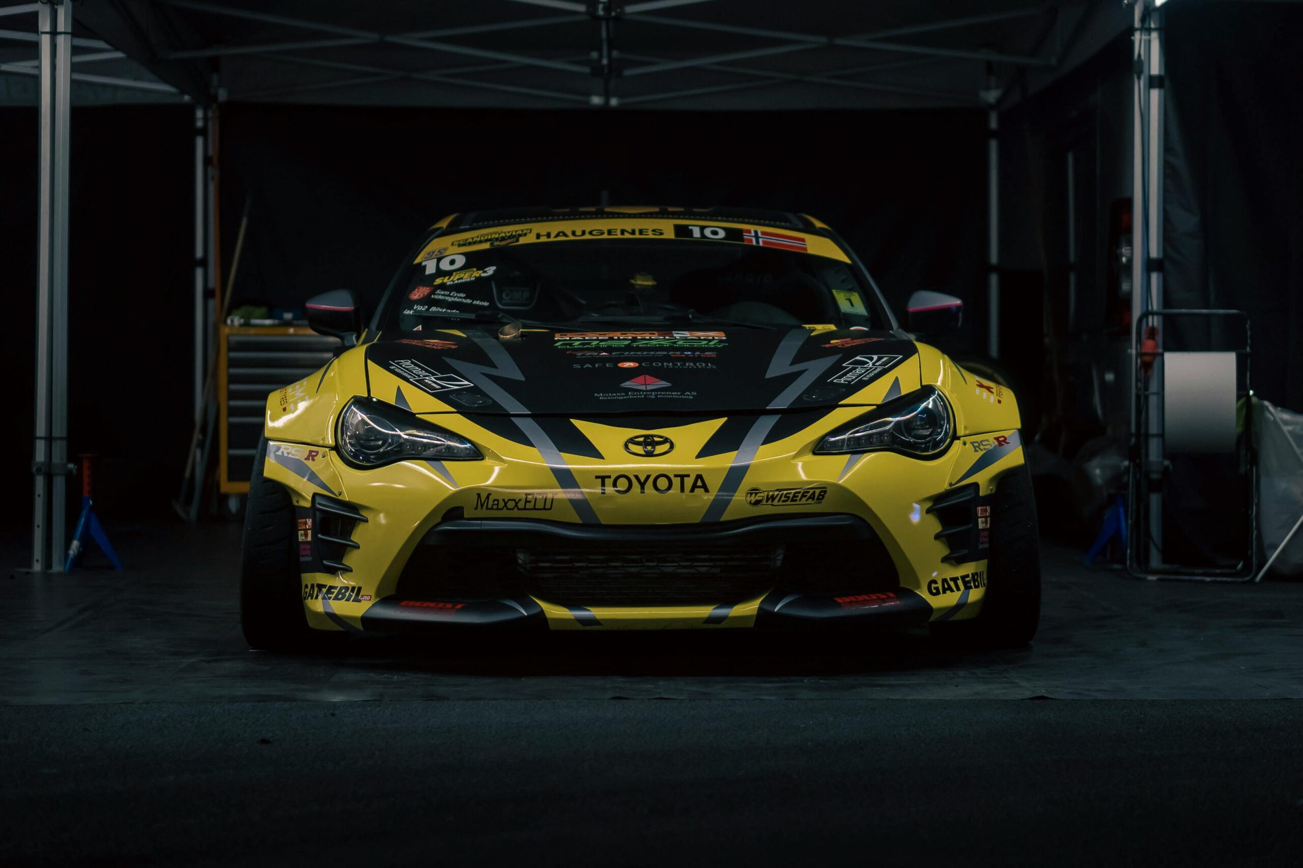 Front view of a yellow Toyota sports car with Norwegian flag in a dimly lit racing garage.
